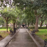 Historic walkway in Bienville Square, Mobile, AL, lined with park benches and massive moss-draped Oak trees leading to a central fountain.